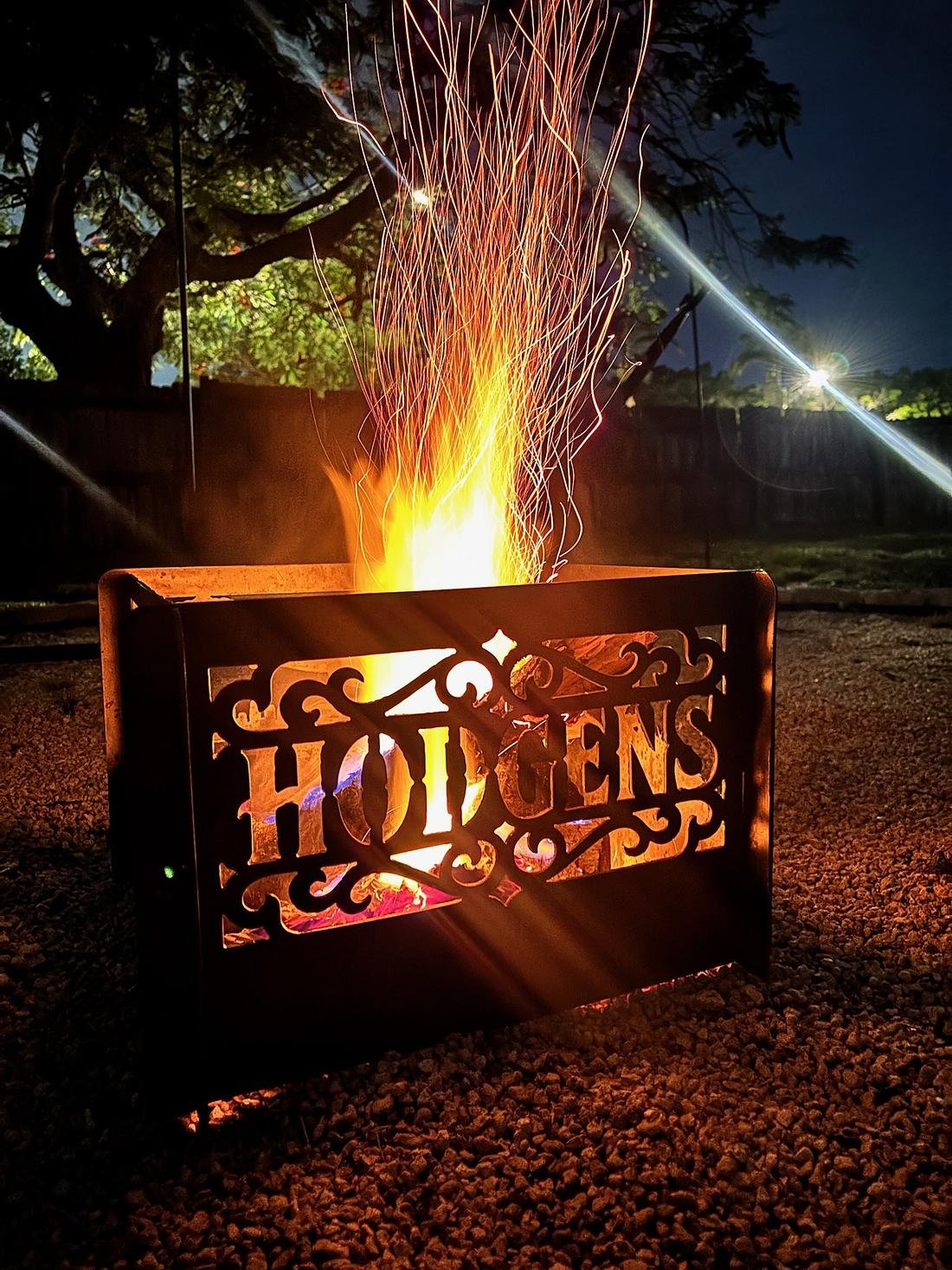 A custom steel fire pit throwing a shower of sparks into the night, a family name cut through the front panel